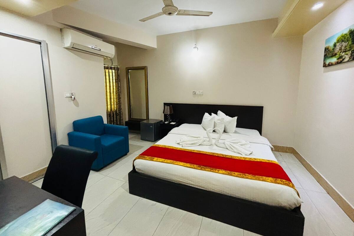A hotel room with a bed, a desk, a chair, a ceiling fan, an air conditioning unit, a mirror, and a painting on the wall.