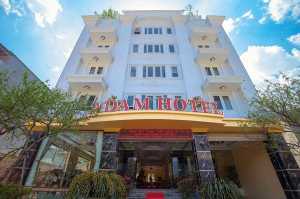 Un hôtel avec une enseigne bien visible portant l’inscription « DAMHOTEL » et une grande entrée surmontée d’un tapis rouge.