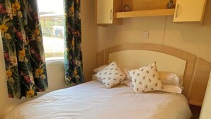 Uma cama com colcha branca e almofadas estampadas, cabeceira de madeira e janela com cortinas florais.