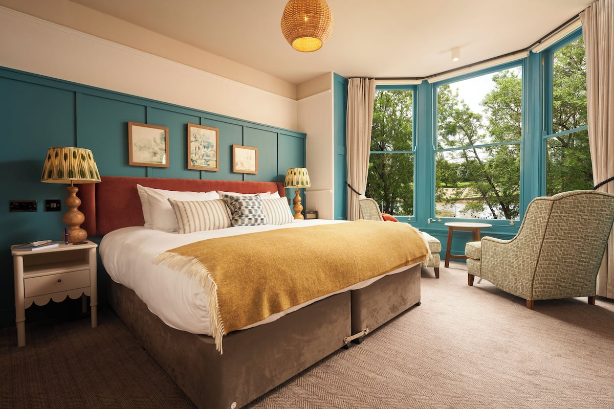 A bedroom with a large bed, a nightstand, a chair, and a view of trees through a window.