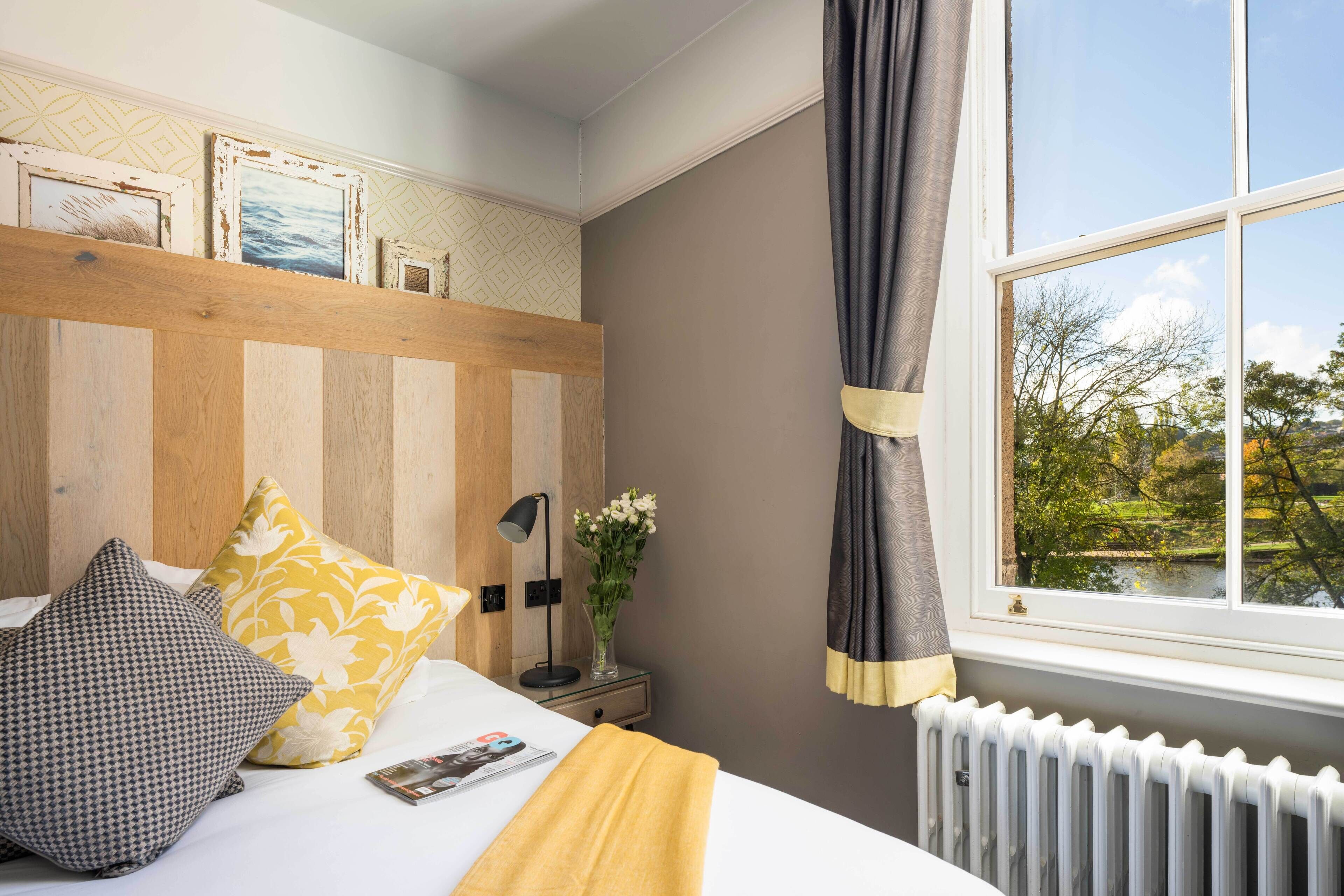 A bedroom with a wooden headboard, a bedside table with a lamp and flowers, a window with curtains, and a view of greenery outside.