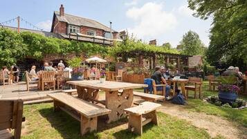 An outdoor seating area with wooden tables and benches, surrounded by greenery and a brick building.
