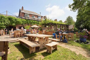 An outdoor seating area with wooden tables and benches, surrounded by greenery and a brick building.