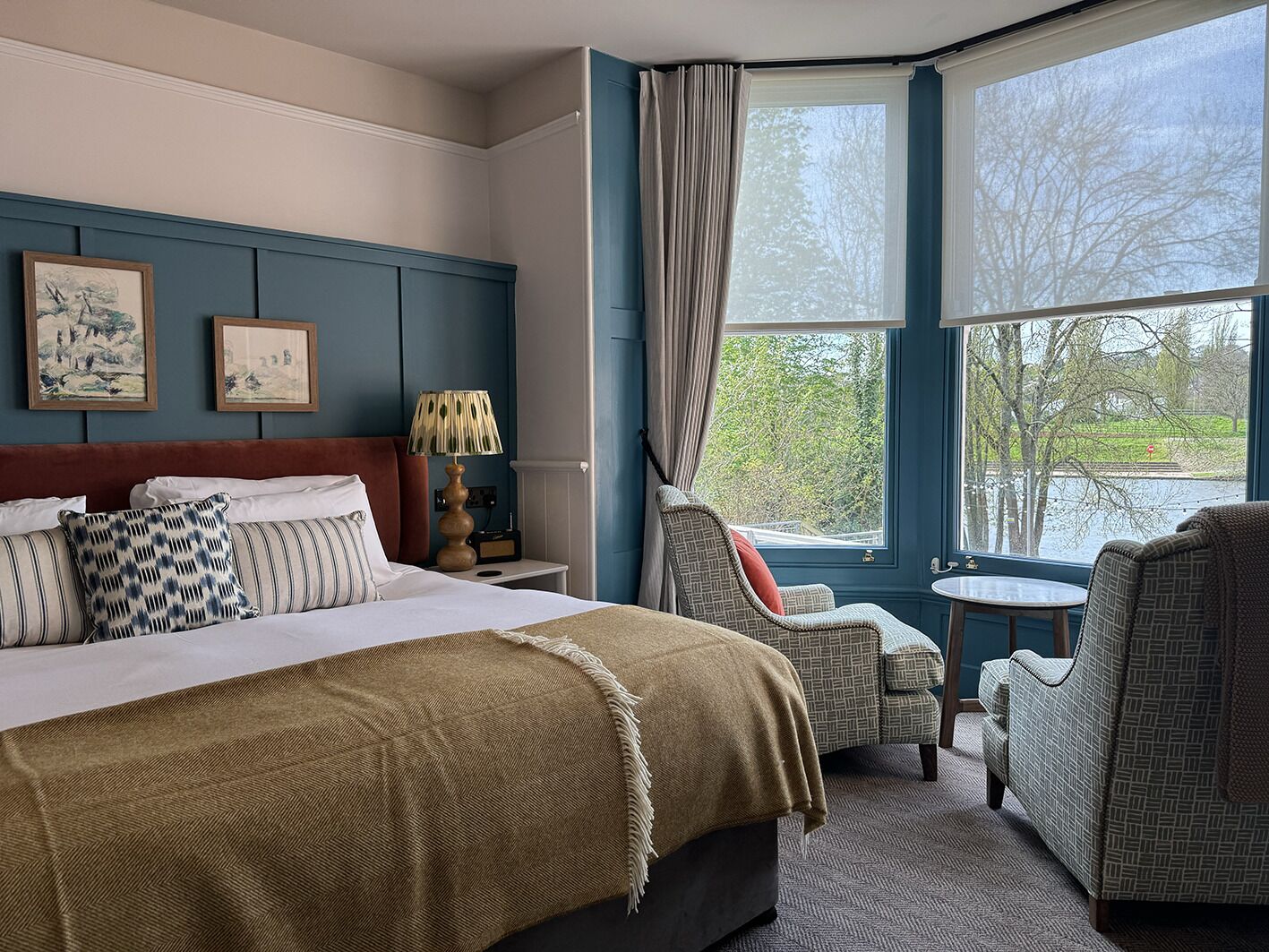 A hotel room with a large bed, two chairs, a small table, and a window with a view of trees and a body of water.