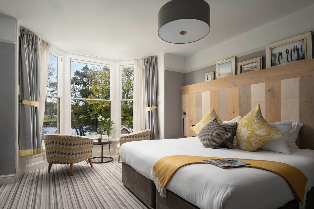 A bedroom with a large bed, a wooden headboard, a window with curtains, a small table, and a chair.