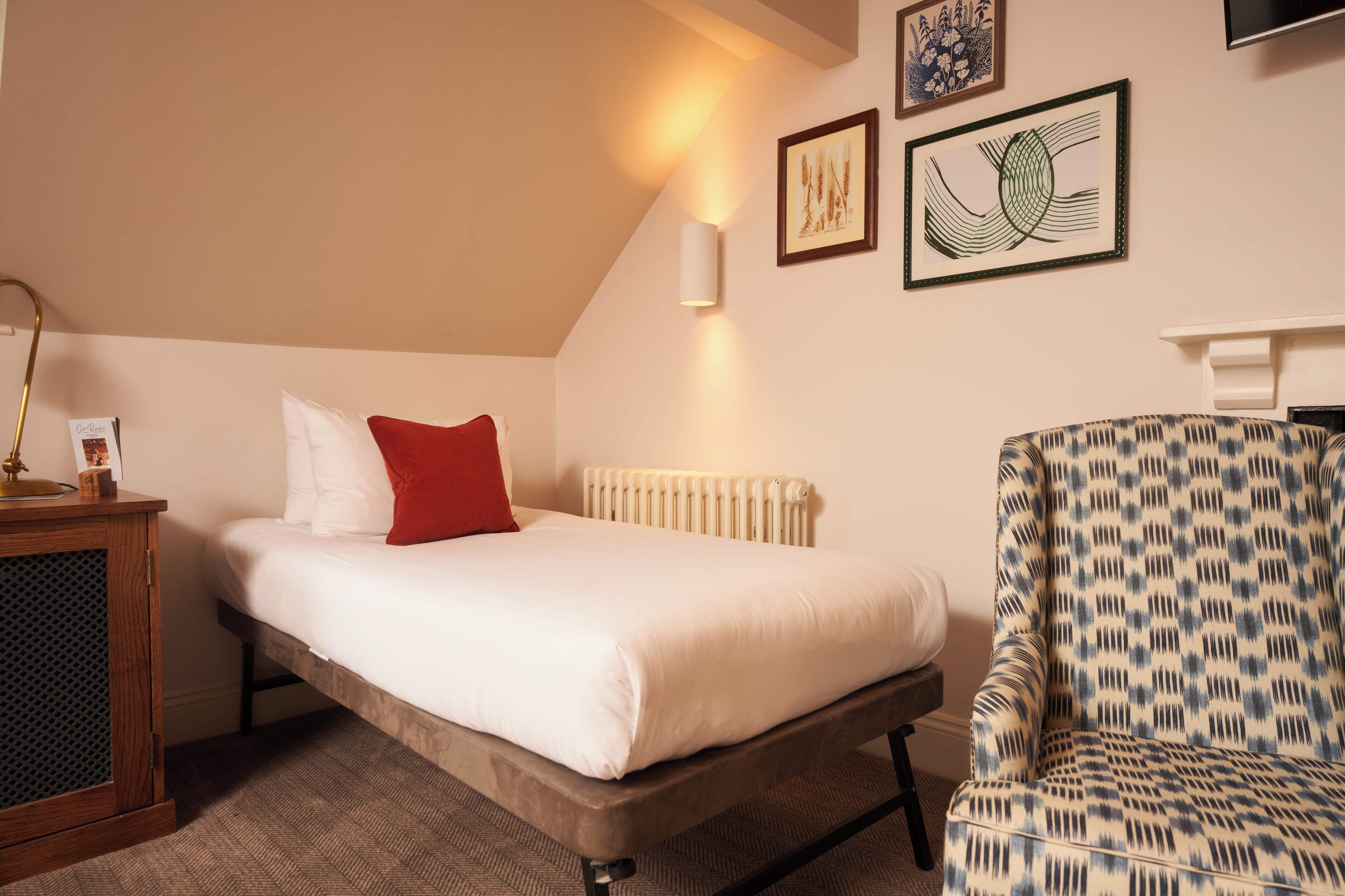 Une chambre avec un lit simple, un oreiller rouge, une table de chevet en bois, un fauteuil à motifs et un radiateur.