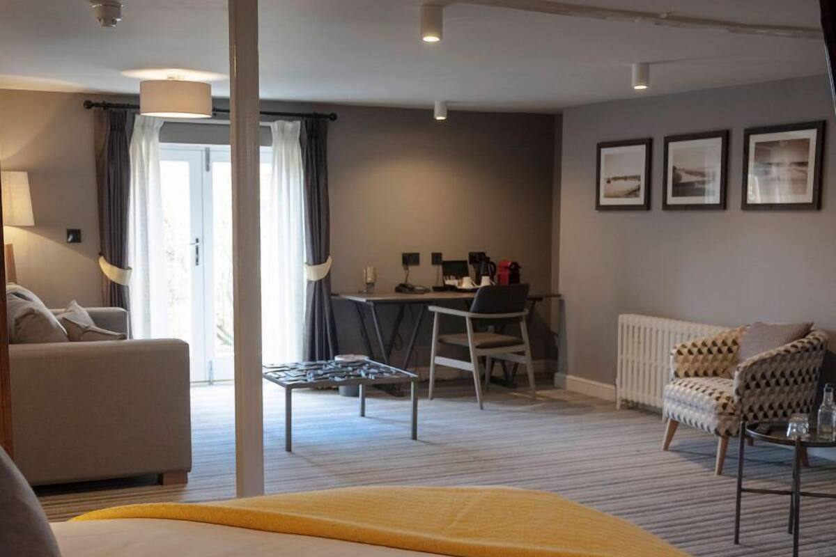 A hotel room with a bed, a desk, a sofa, and a chair. There is a window with curtains, a radiator, and framed pictures on the wall.