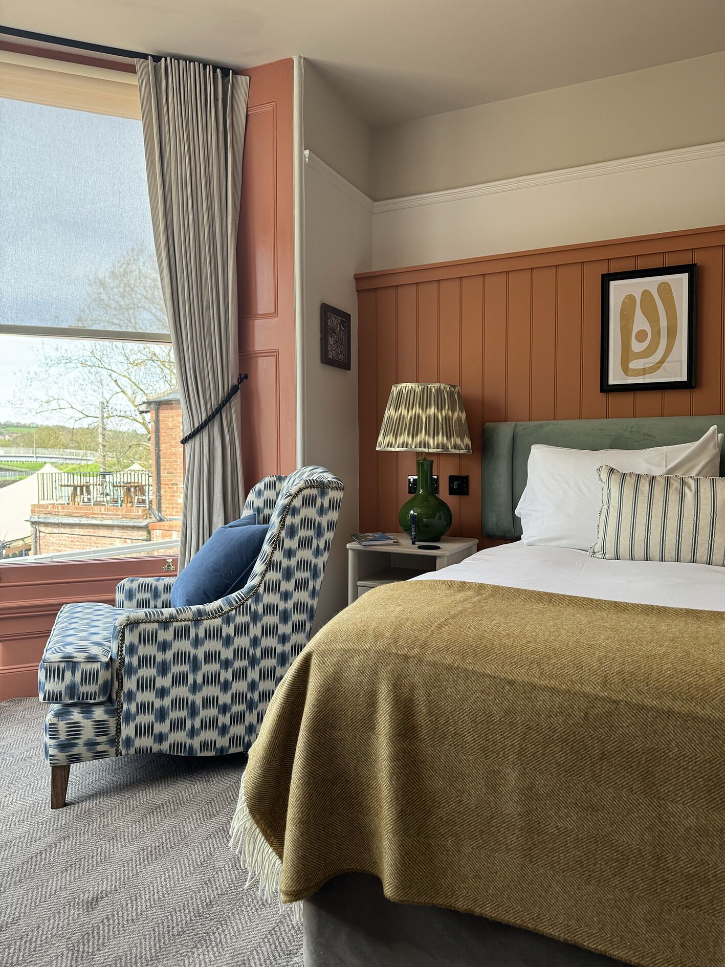 A hotel room with a bed, a chair, a nightstand, a lamp, and a view of the outdoors.