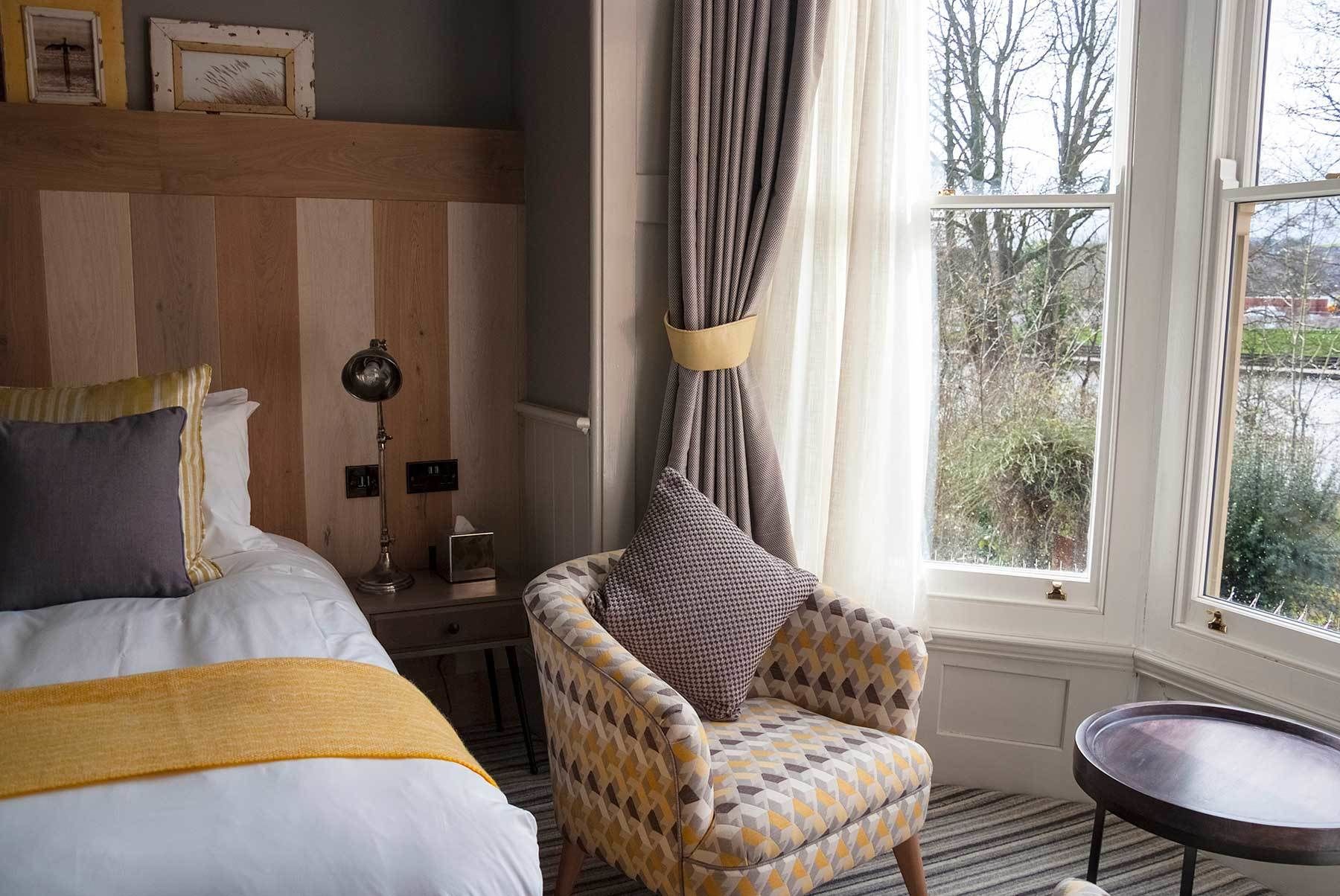 Habitación de hotel con cama, sillón, mesita de noche, lámpara y ventana con vistas a árboles.