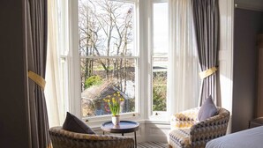 A hotel room with a large window, two patterned armchairs, a small round table with a vase of flowers, and a bed with a white duvet. - Mill on the Exe (Exeter)