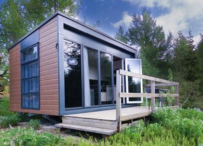 Una cabaña moderna con amplios ventanales, una terraza de madera y un bosque circundante.