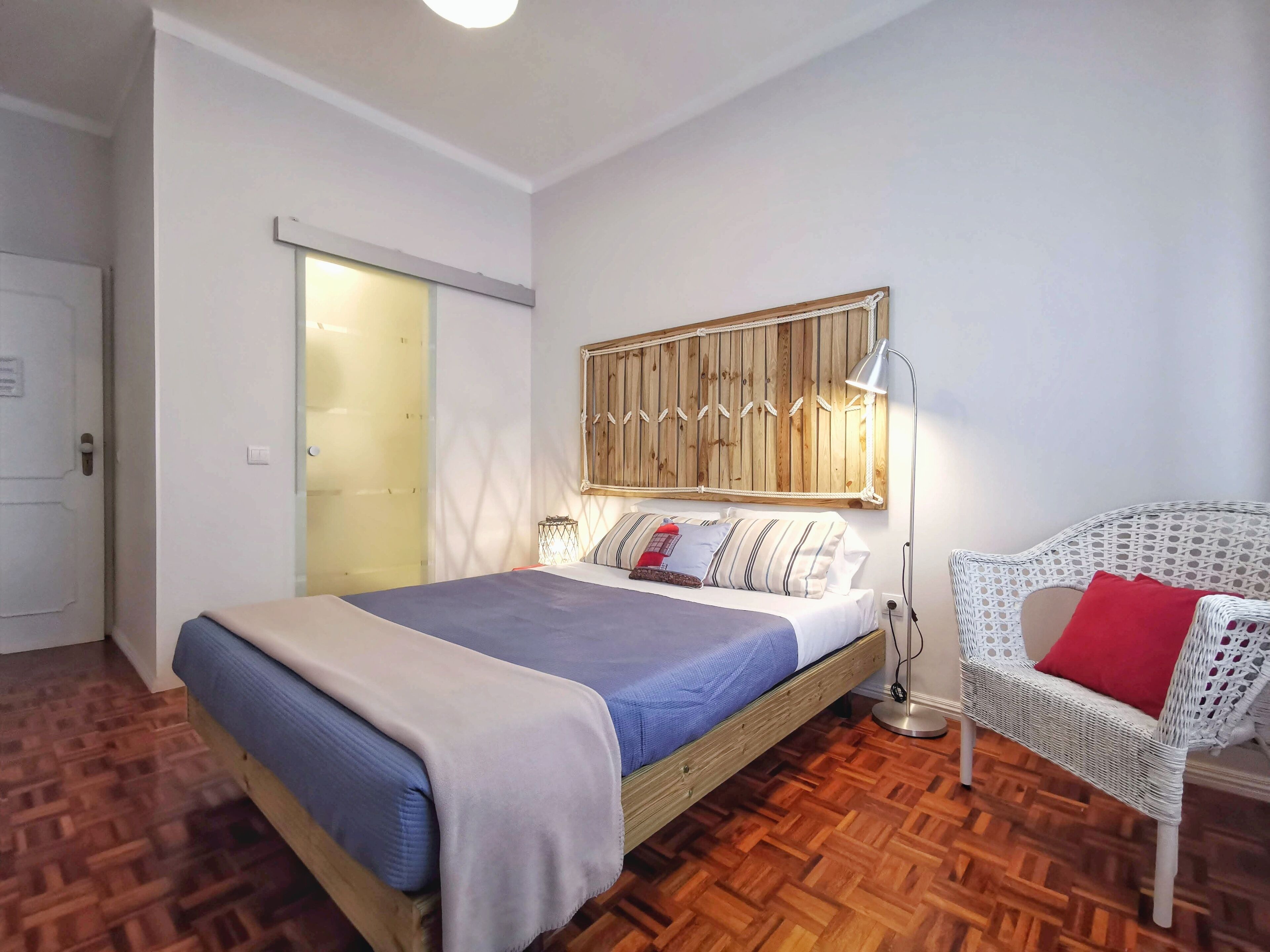 A bedroom with a bed, a white wicker chair, a wooden headboard, and a wooden floor.