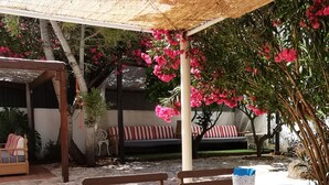 Uma área de refeições ao ar livre com mesas e cadeiras de madeira, um espaço coberto para sentar e um jardim com flores cor-de-rosa.