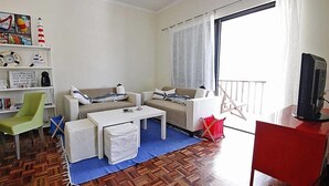 Sala de estar com sofá, mesa de centro e TV em um suporte. Estante com livros e pequena mesa com abajur. Janela com cortinas visível.
