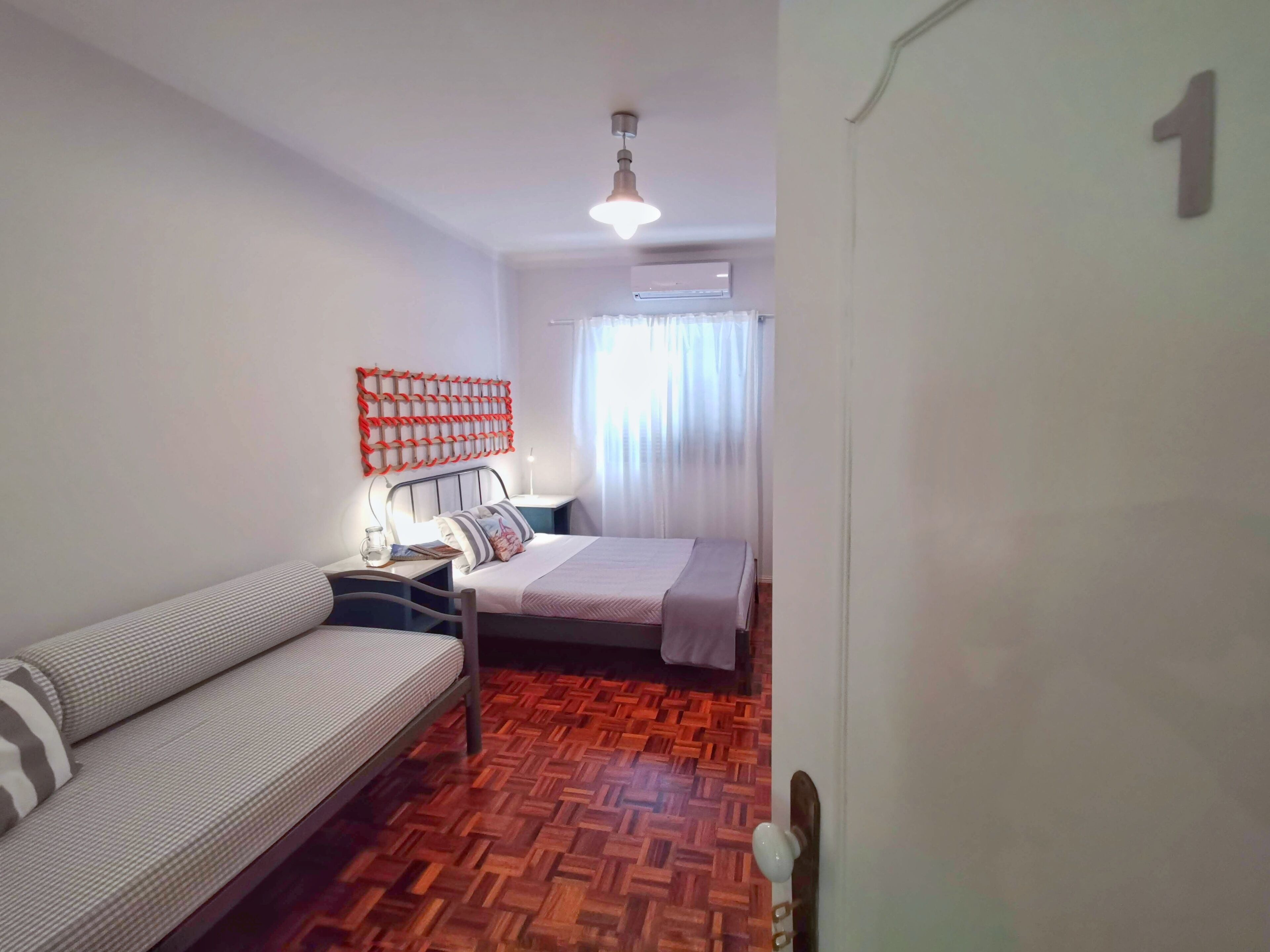 A room with a bed, a sofa, a wall-mounted light fixture, and a patterned wooden floor.