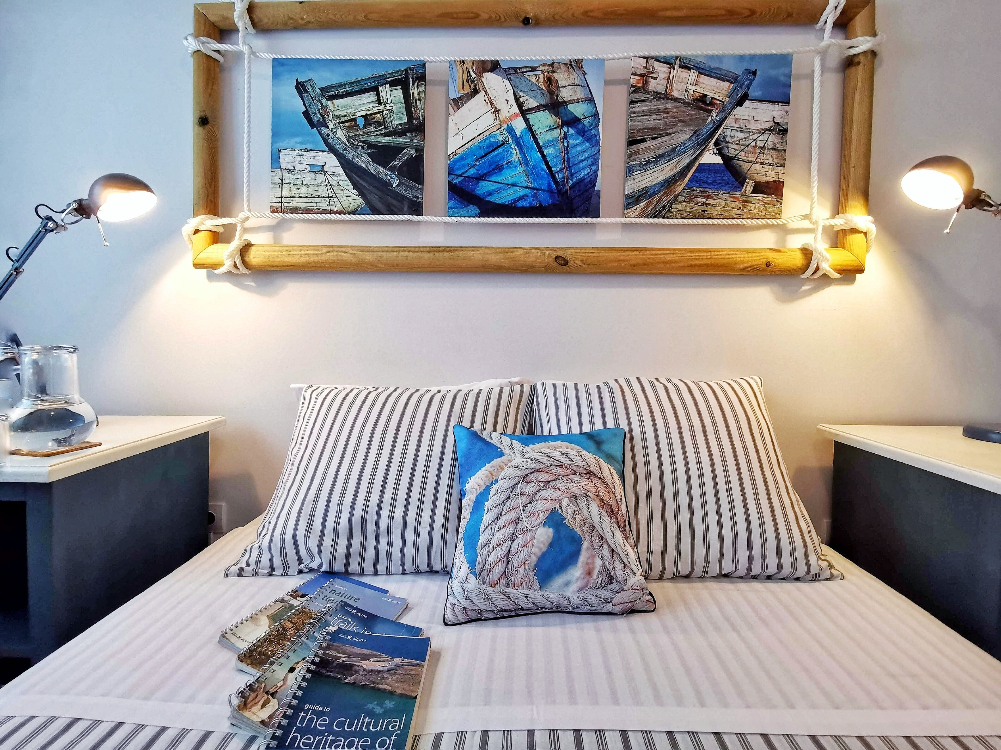 Quarto bem arrumado com uma cama, almofadas listradas, uma mesinha com abajur e um quadro na parede de barcos.