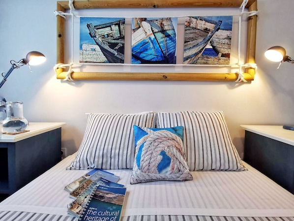 A neatly arranged bedroom with a bed, striped pillows, a small table with a lamp, and a wall-mounted painting of boats. - Roaz de Bandeira House (Olhao)