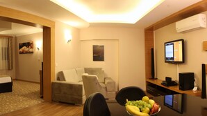 A hotel room with a sofa, a dining table with fruit, a TV, and a desk with a computer. - Grand Zeybek Hotel (Izmir)