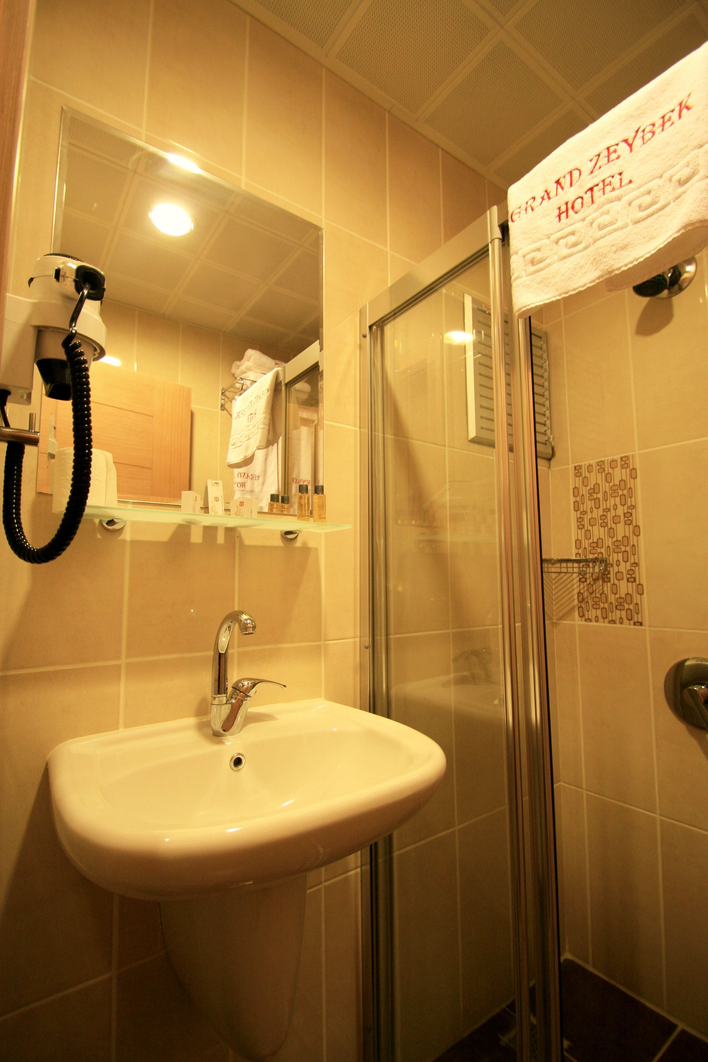 A bathroom with a sink, mirror, and shower. A towel hangs on the wall, and a sign reads "Grand Zeybek Hotel".