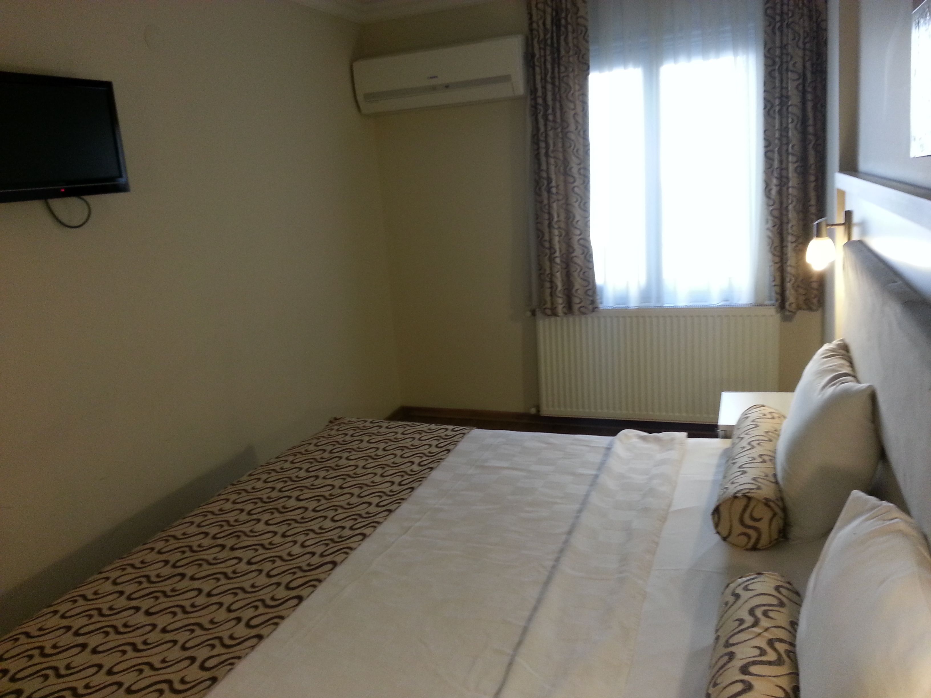 A hotel room with a bed, a flat-screen TV mounted on the wall, an air conditioning unit, and a window with curtains.