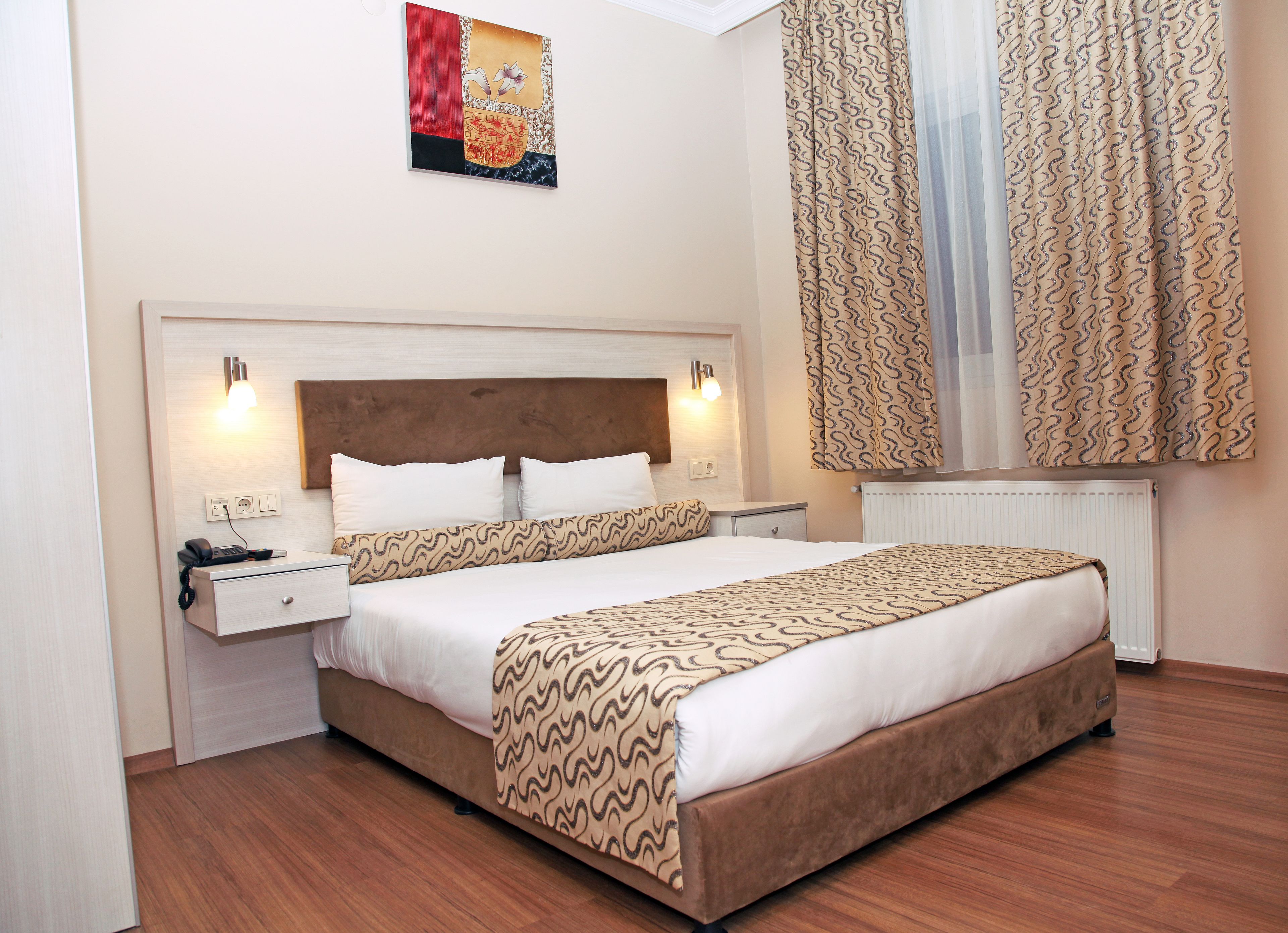 A hotel room with a bed, bedside tables, a nightstand, a wall-mounted artwork, and patterned curtains.