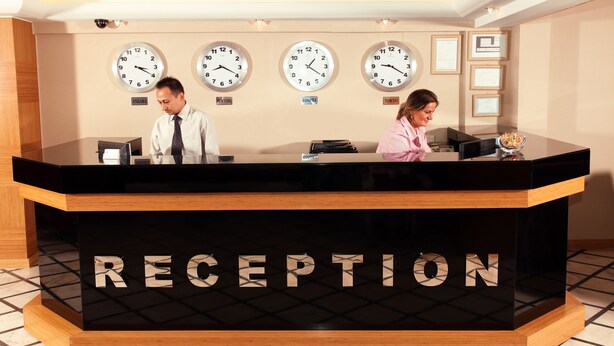 A reception desk with clocks displaying different cities, a nameplate reading "RECEPTION," and a tiled floor. - Grand Zeybek Hotel (Izmir)