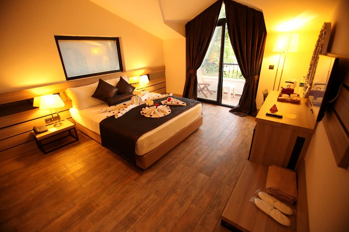 Habitación de hotel con una cama grande, mesitas de noche con lámparas, suelo de madera, un balcón con vistas y un escritorio con espejo y lámpara.