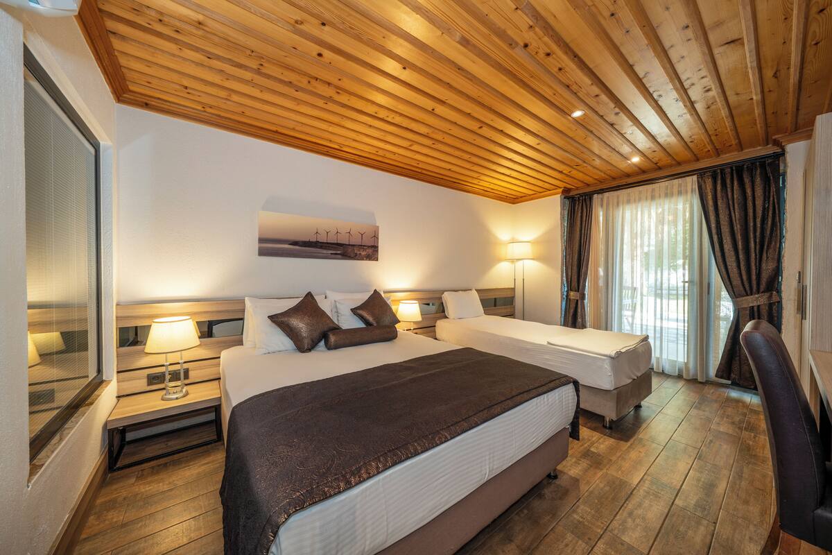 Una habitación de hotel con techo de madera, dos camas, una mesita de noche con lámpara, una silla y una ventana con cortinas.