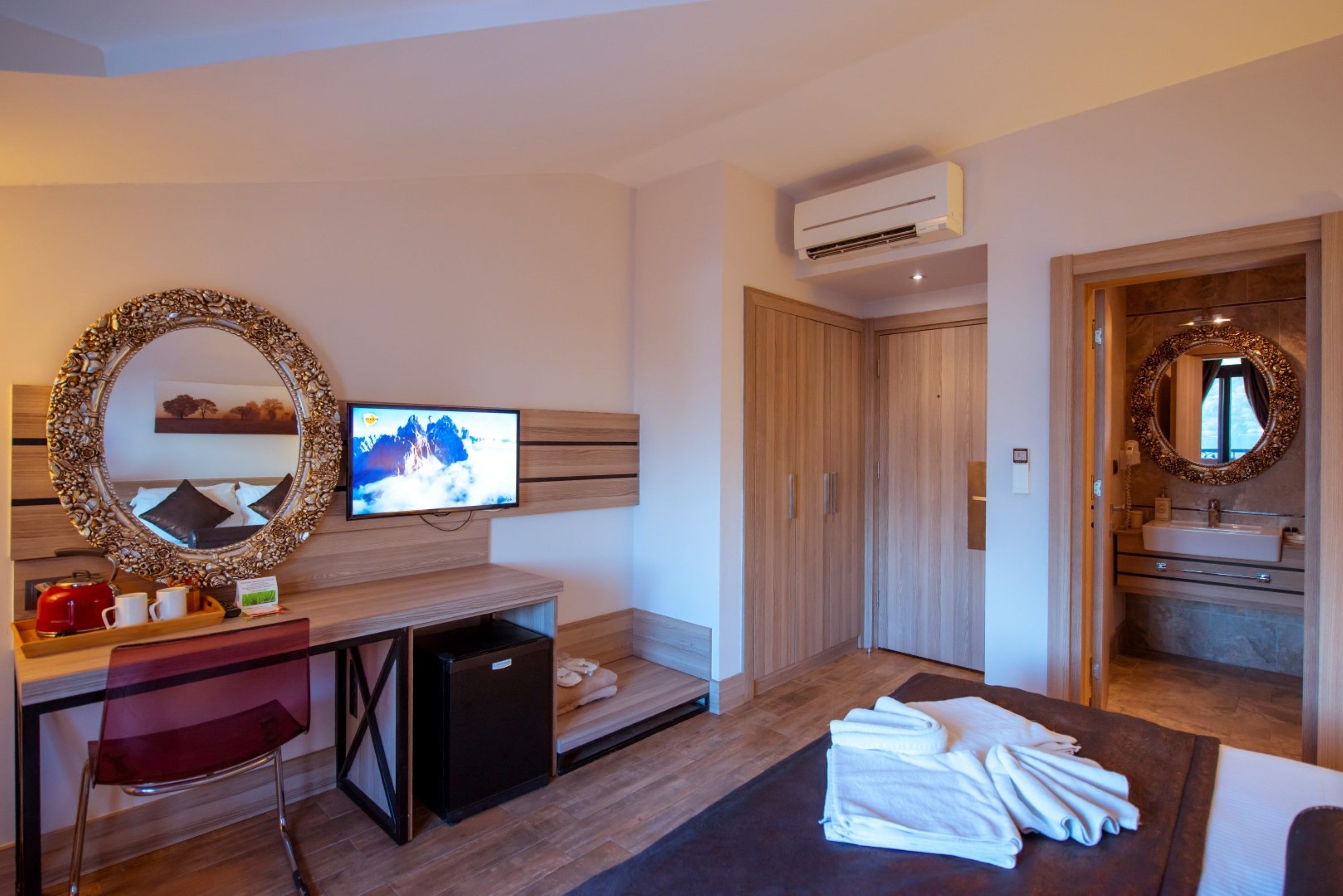 Una camera d'albergo moderna con una scrivania in legno, una televisione, uno specchio, un piccolo frigorifero e un bagno con lavandino e specchio.