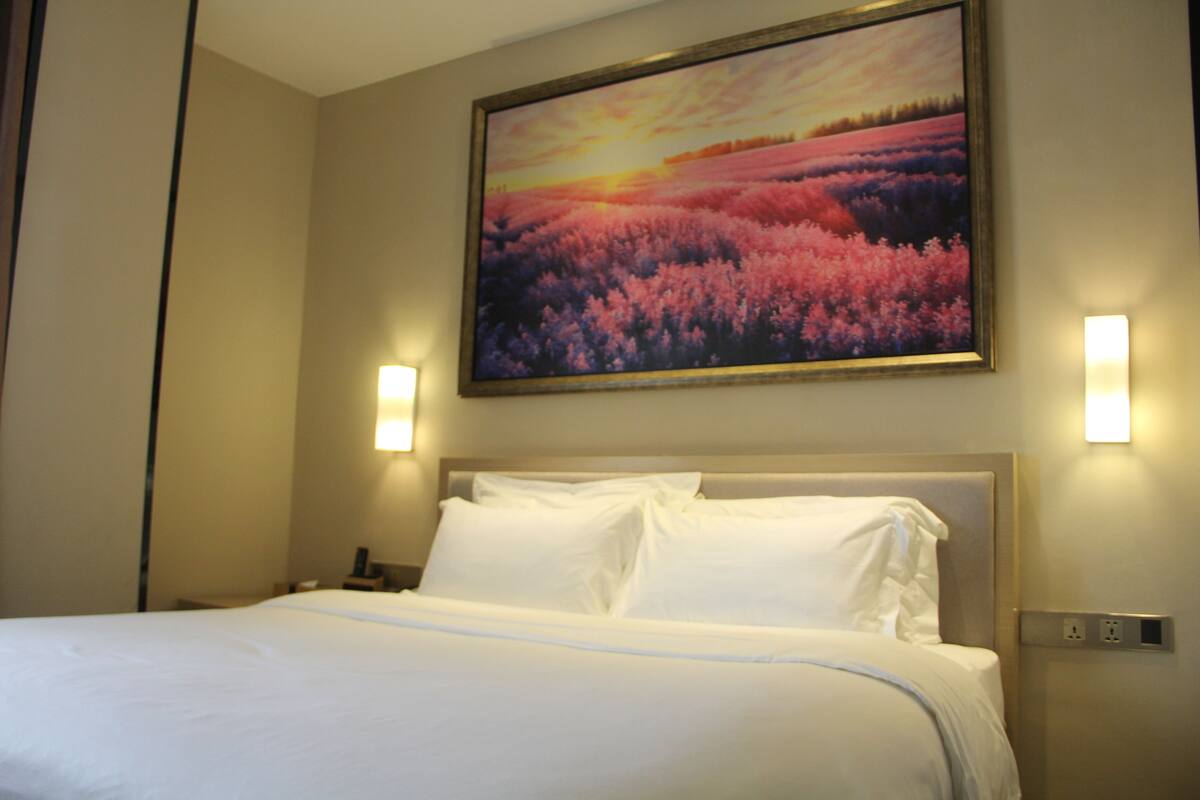 A hotel room with a large bed, two bedside lamps, and a painting of a sunset over a field.