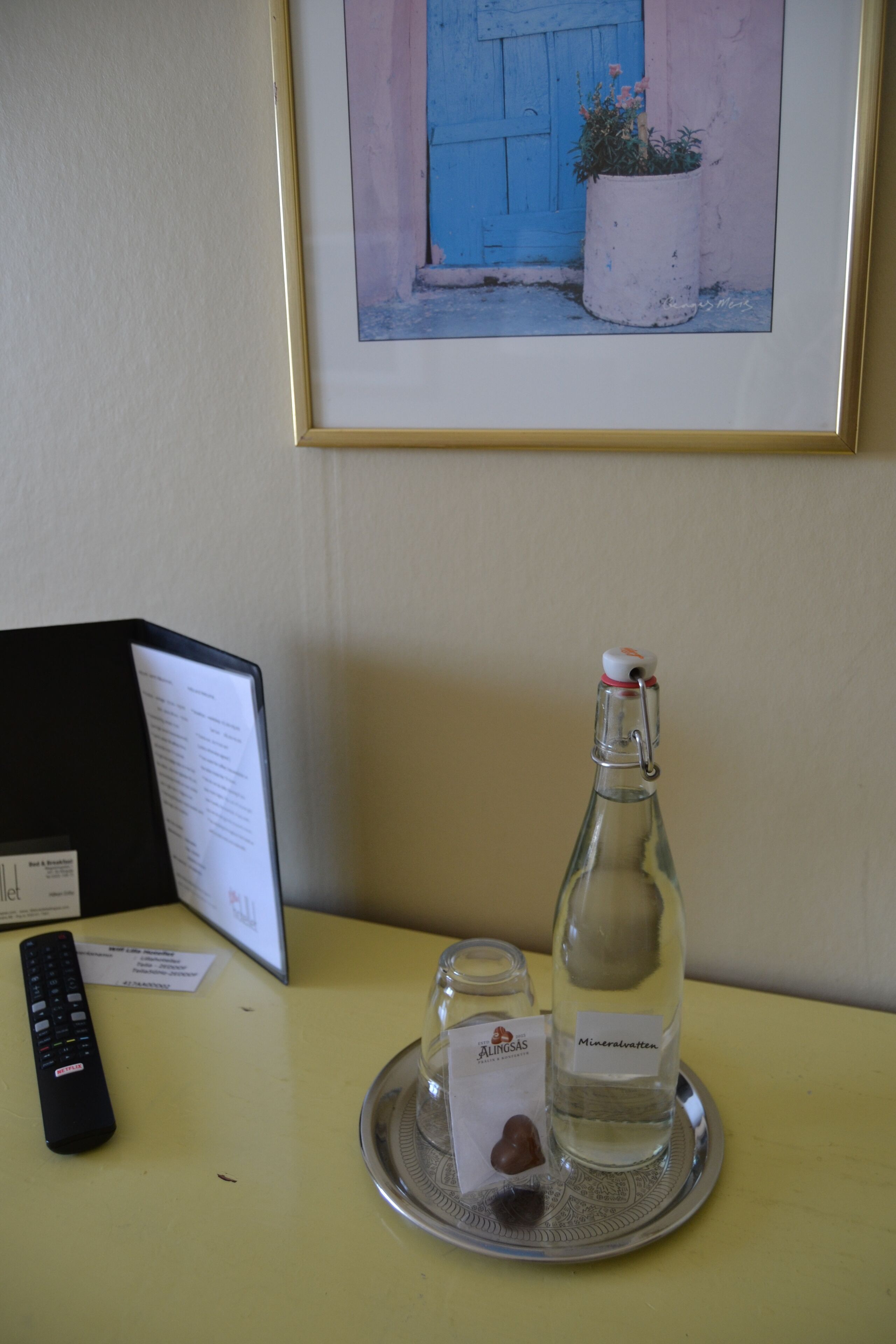 Una mesa con un control remoto, una botella de agua mineral, un vaso pequeño y un marco con una foto de una puerta azul y una planta en maceta.