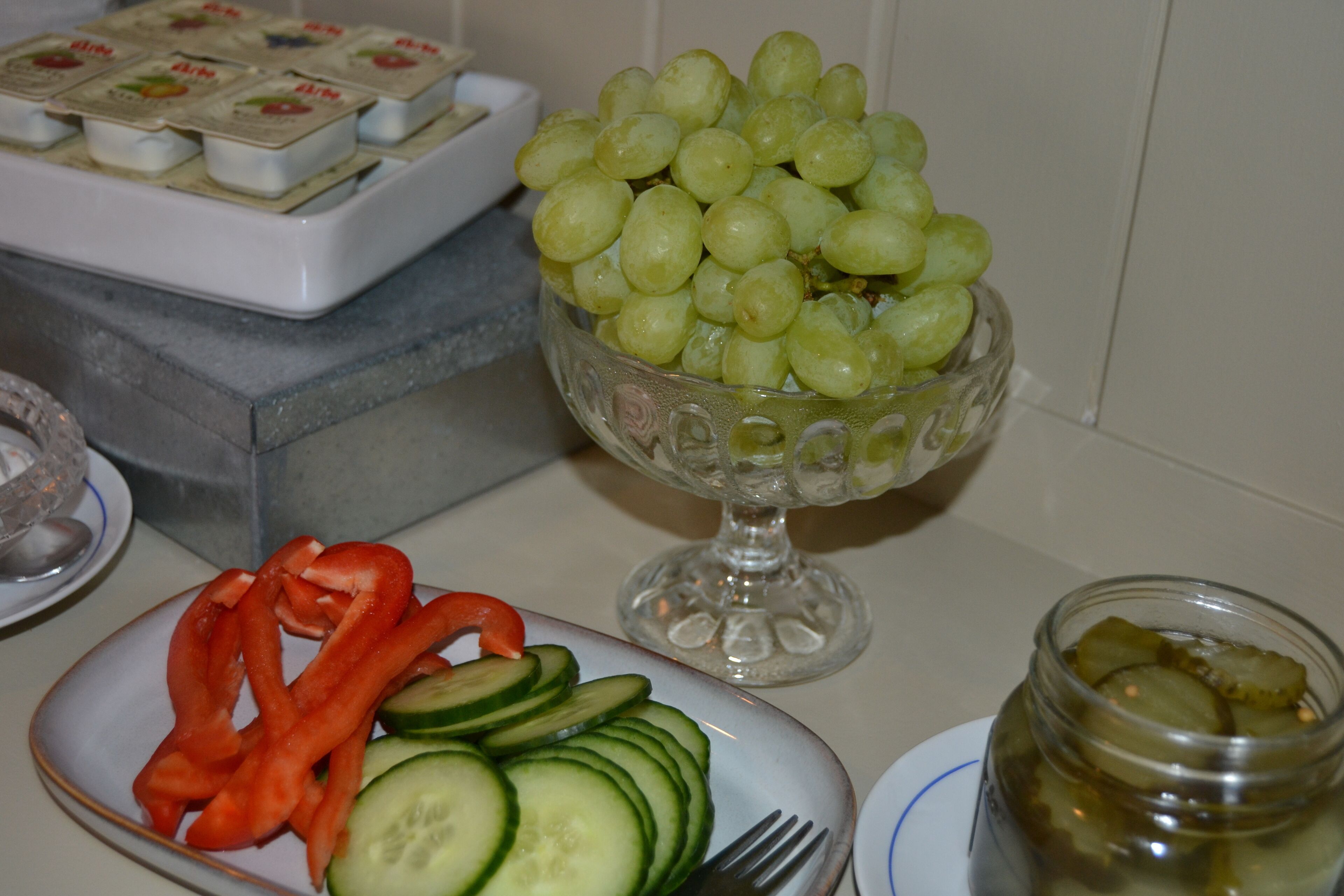 A table with a bowl of green grapes, a plate with sliced cucumbers and red bell peppers, and a jar of pickles.