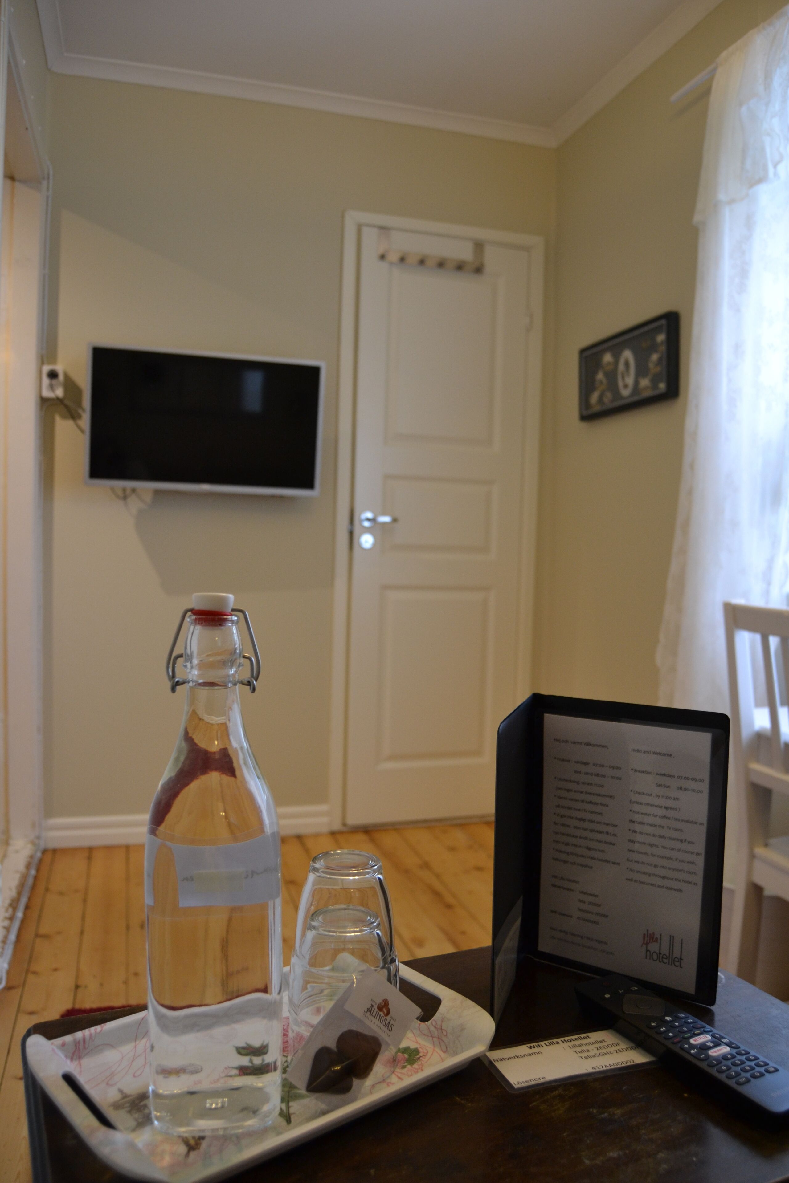 A hotel room with a wooden floor, a television mounted on the wall, a bottle of wine, a glass, a tray with a card, and a remote control.