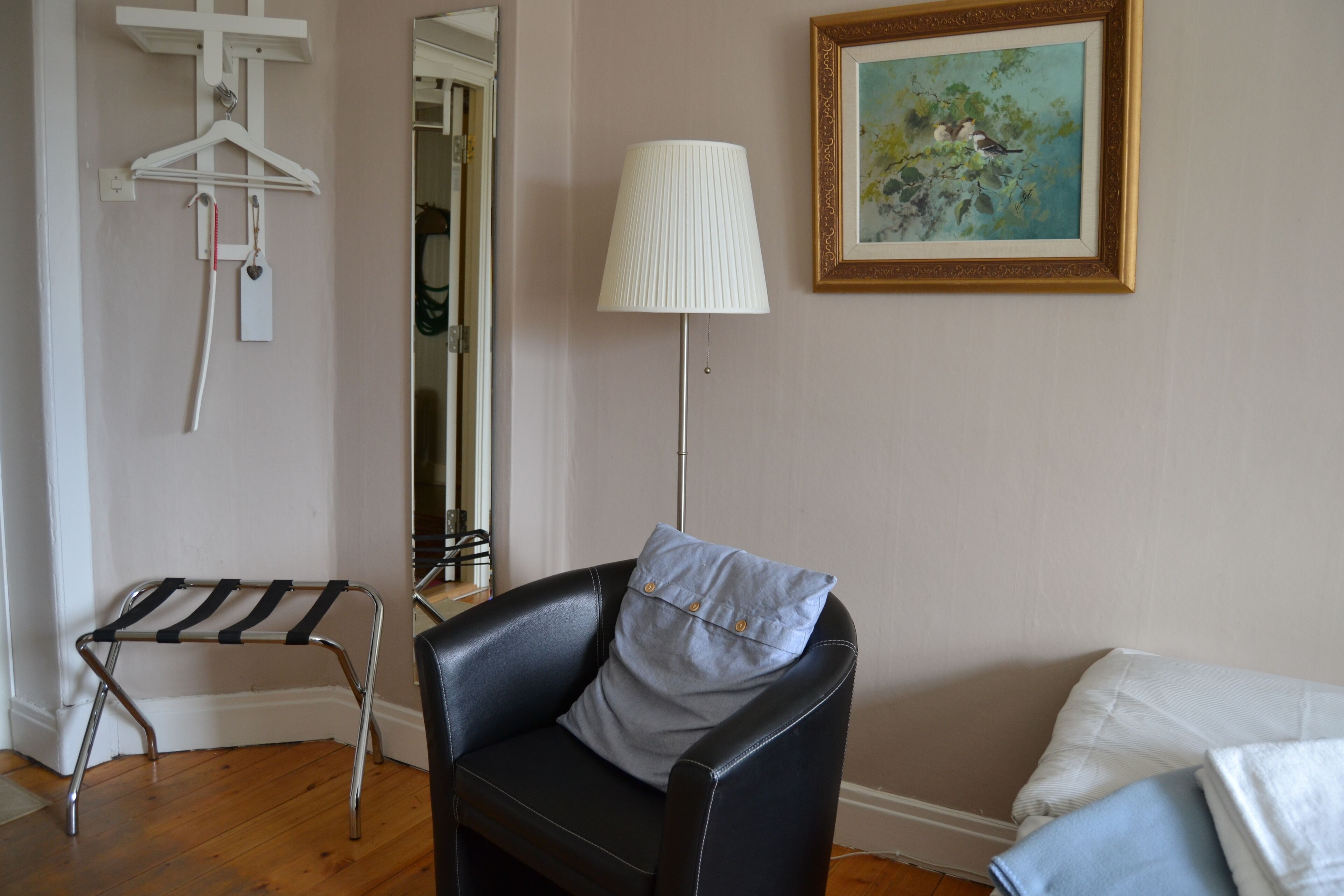 Una habitación con una butaca de cuero negro, un sillón blanco, una lámpara de pie, un cuadro enmarcado y un perchero con ganchos y una etiqueta.