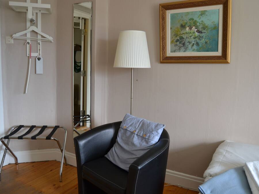 Una habitación con una butaca de cuero negro, un sillón blanco, una lámpara de pie, un cuadro enmarcado y un perchero con ganchos y una etiqueta.