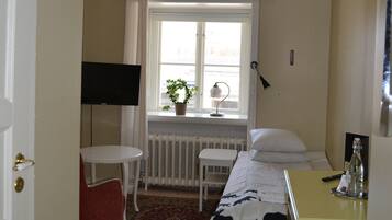 Ett litet hotellrum med en säng, ett litet bord, en stol, ett element, ett fönster med gardiner, en tv som är monterad på väggen och en växt på fönsterbrädan.