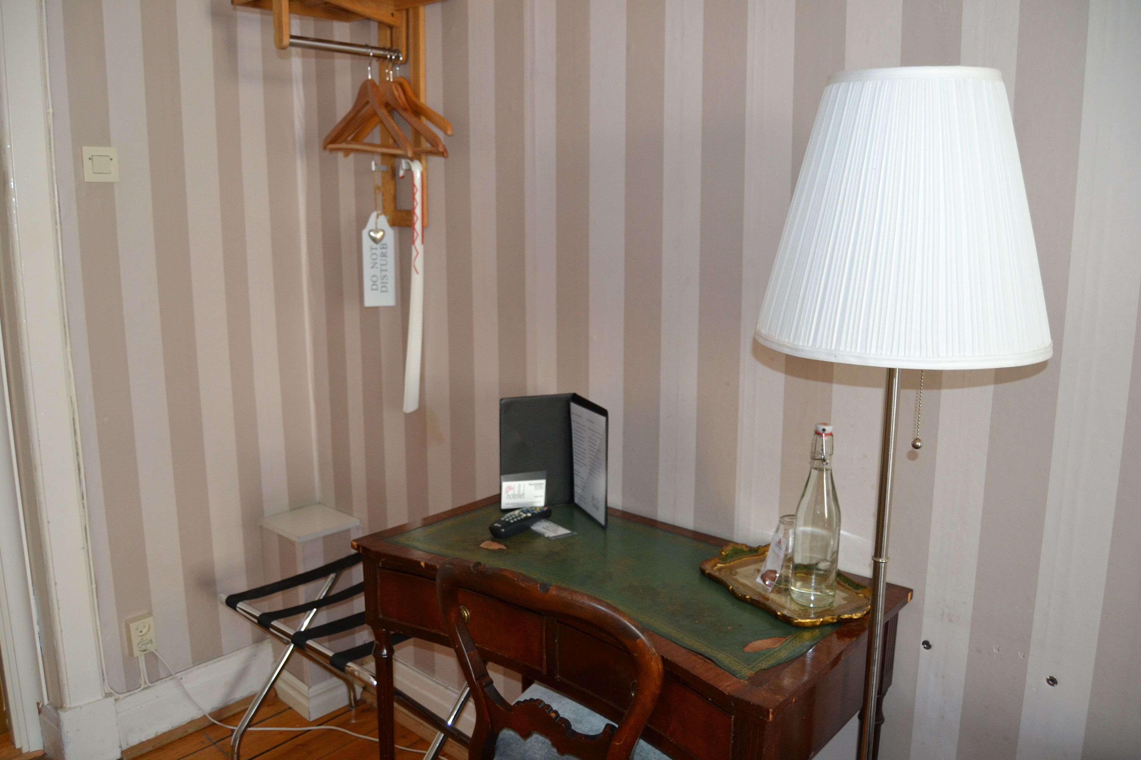 A room with striped wallpaper, a desk with a lamp, a chair, a hanging coat rack, and a small shelf.