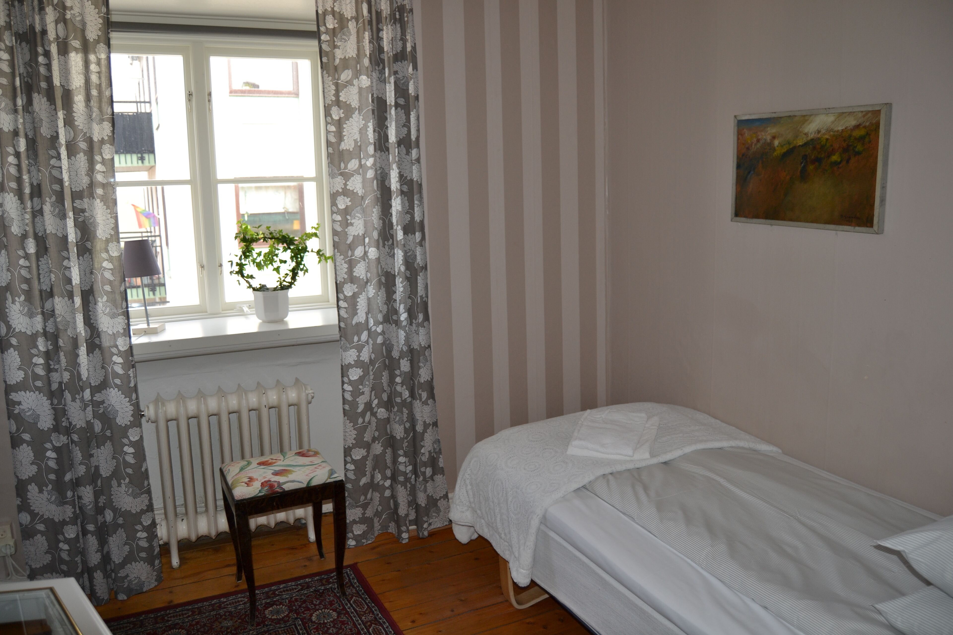 Un dormitorio con una cama individual, un radiador, una ventana con cortinas, una planta en maceta y un cuadro en la pared.