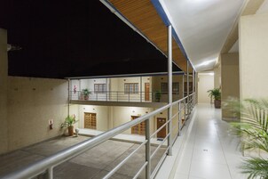 Un pasillo interior de varios niveles con barandillas de metal, plantas en macetas y puertas que dan a habitaciones.