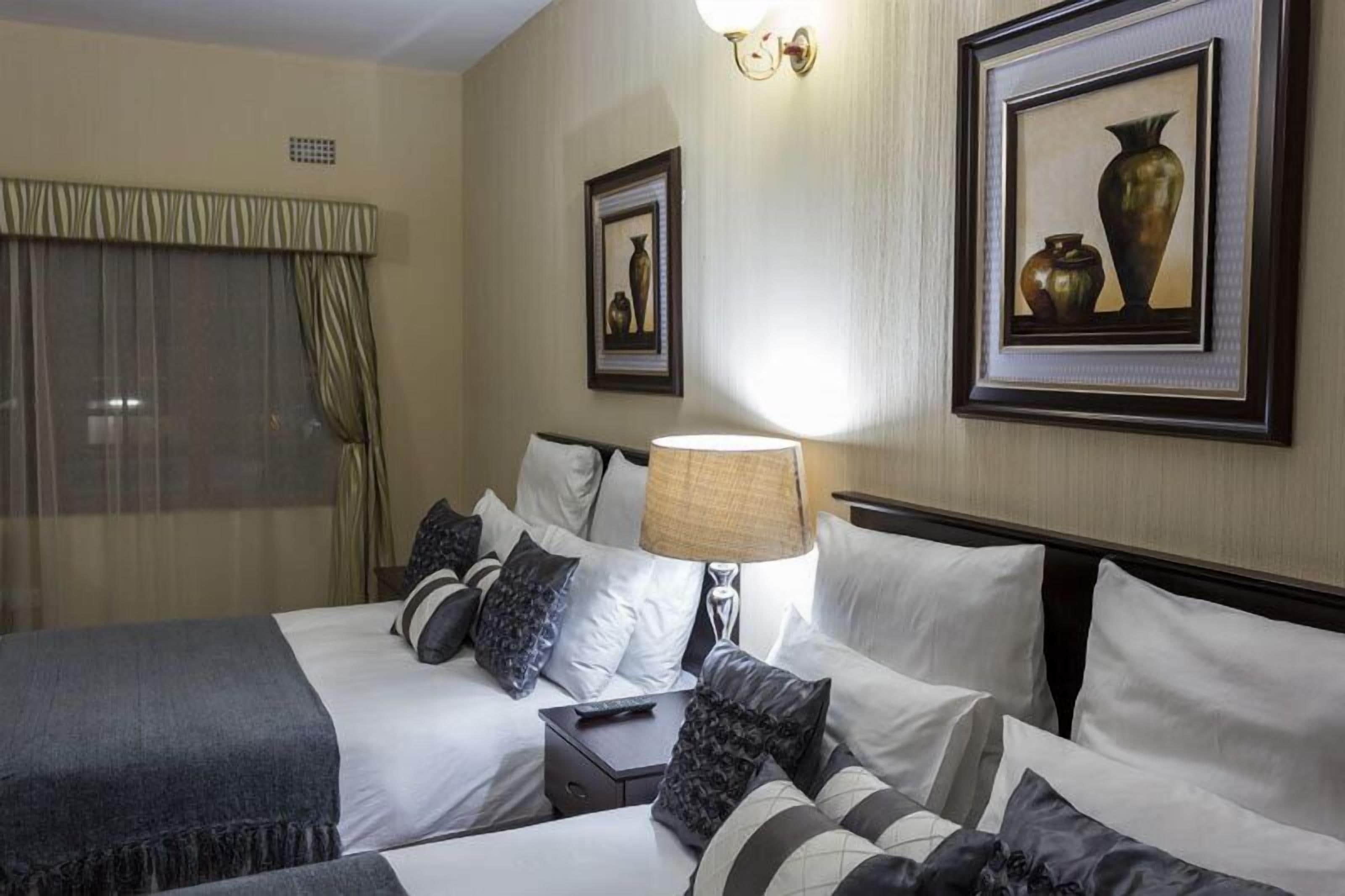 A hotel room with two beds, a bedside table with a lamp, and framed artwork on the walls.