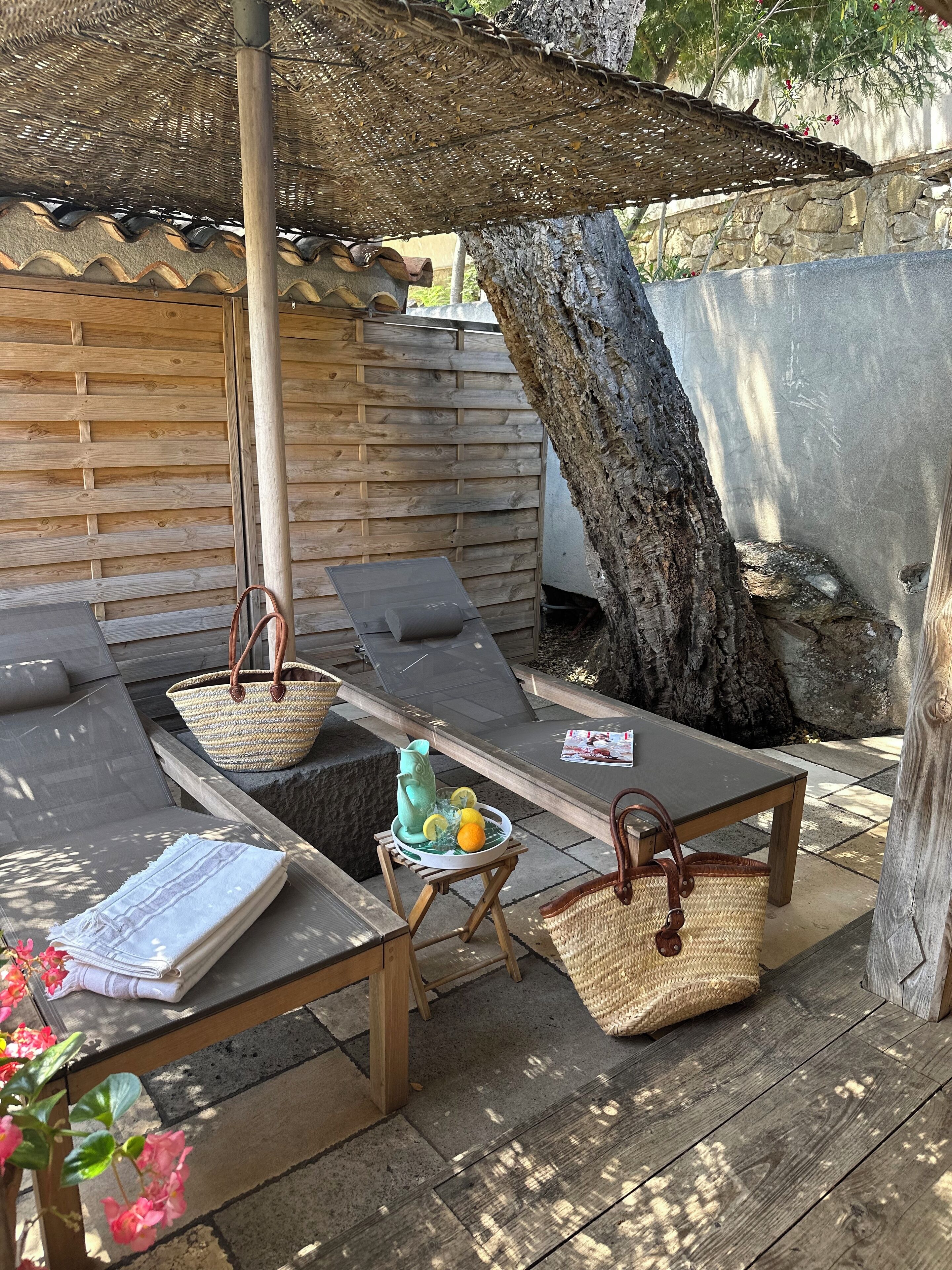 An outdoor seating area with wooden loungers, a table with a basket and a decorative item, and a tree trunk.
