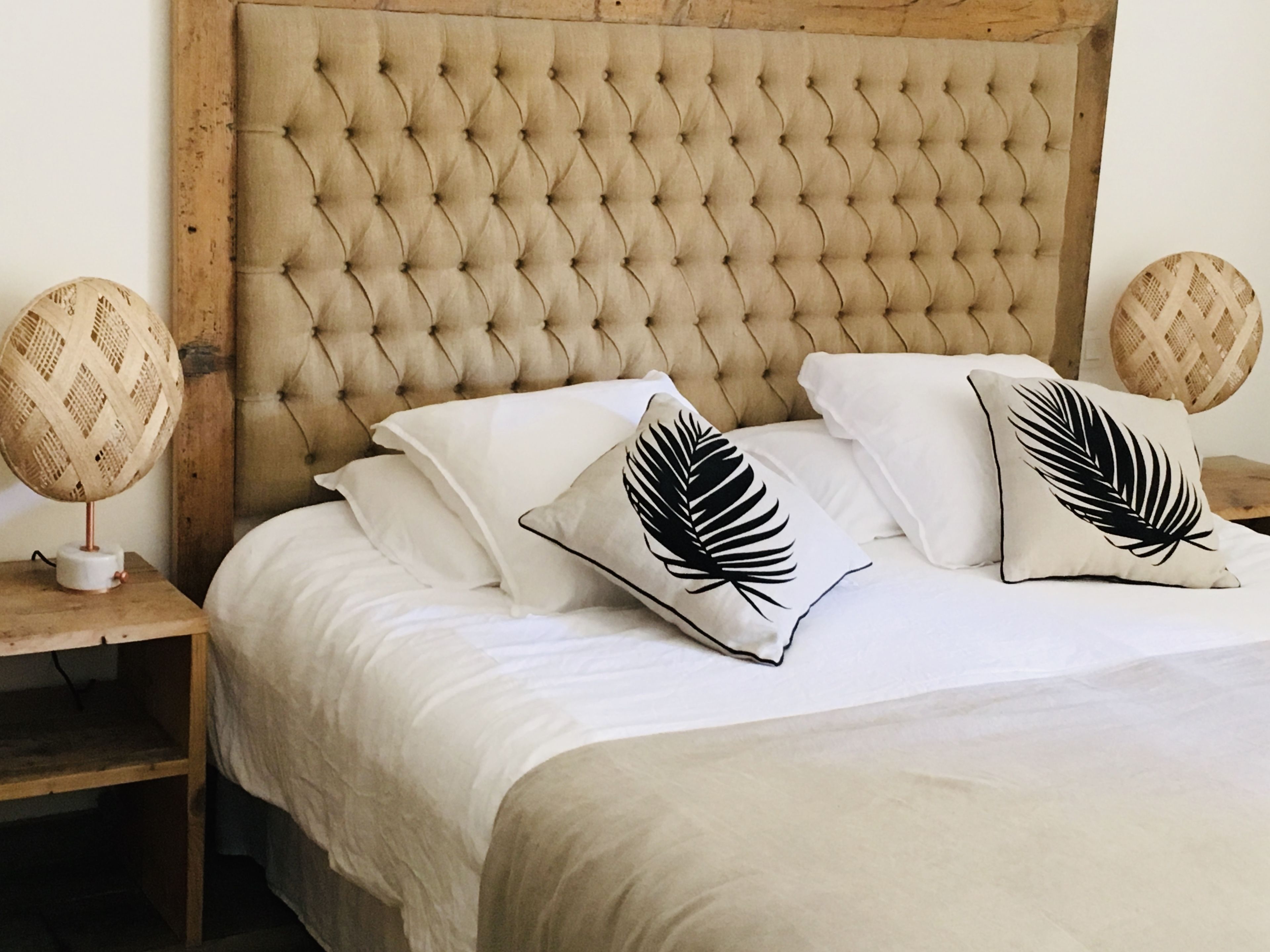 Uma cama com cabeceira estofada, roupa de cama branca e duas almofadas decorativas com padrão de folha.