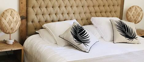 Una cama con cabecera capitonada, ropa de cama blanca y dos almohadones decorativos con diseño de hojas.