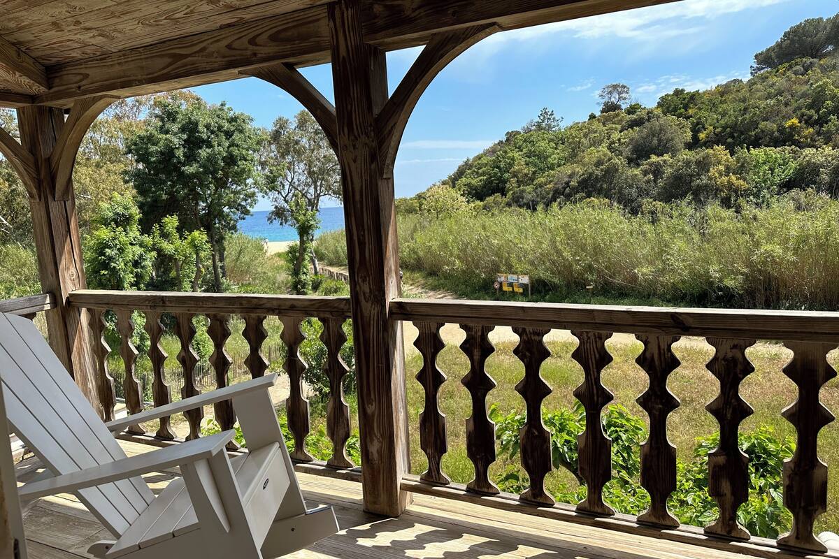 A wooden deck with a white Adirondack chair, overlooking a beach and forested hills.