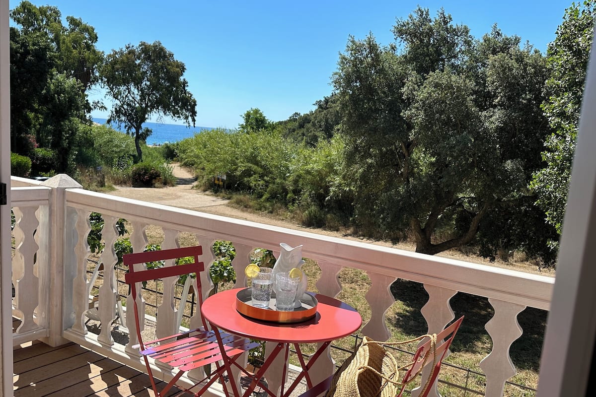 A wooden deck with a table and chairs, overlooking a natural landscape with trees and a view of the sea.