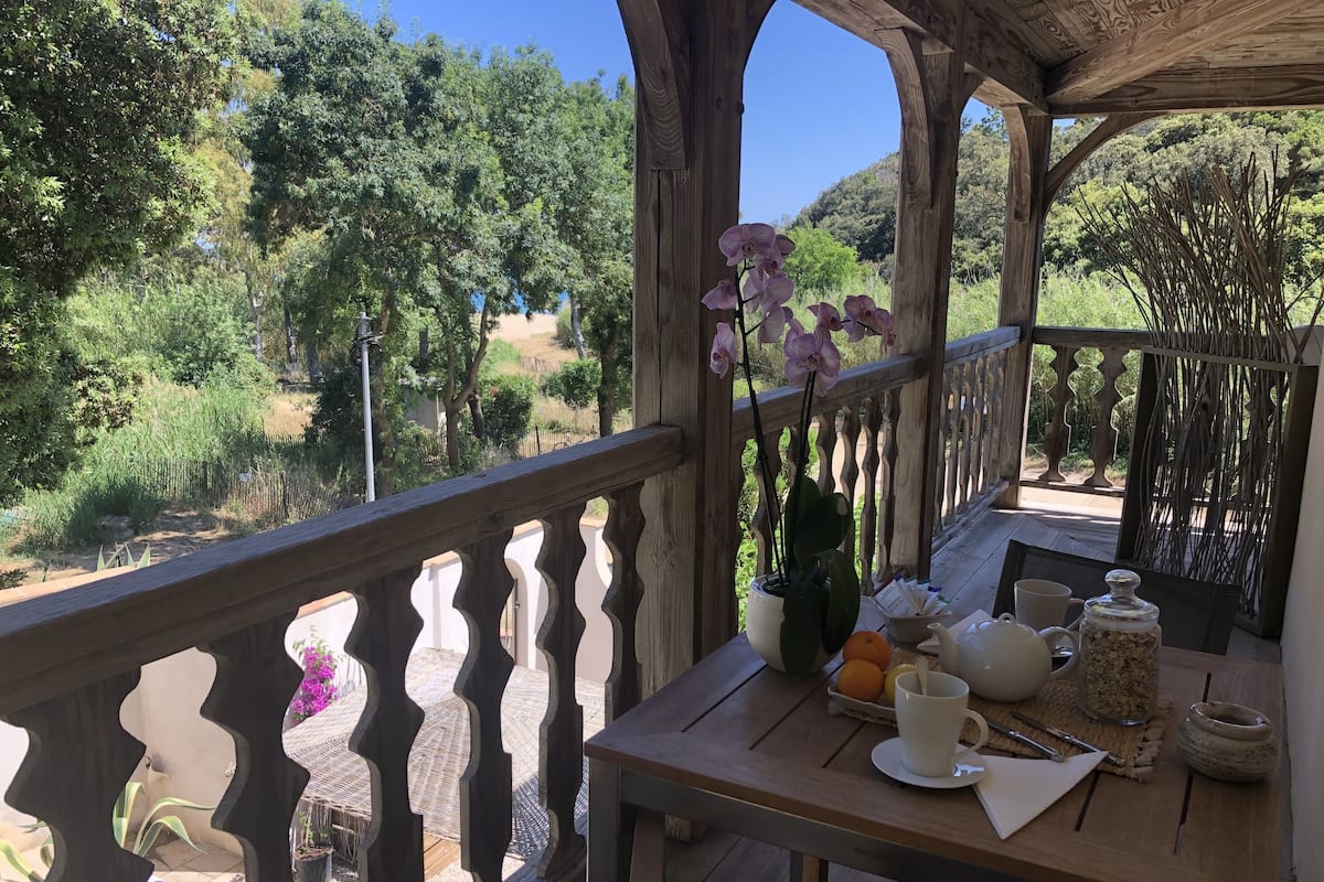 A wooden deck with a table set for breakfast, overlooking a garden with trees and a clear sky.