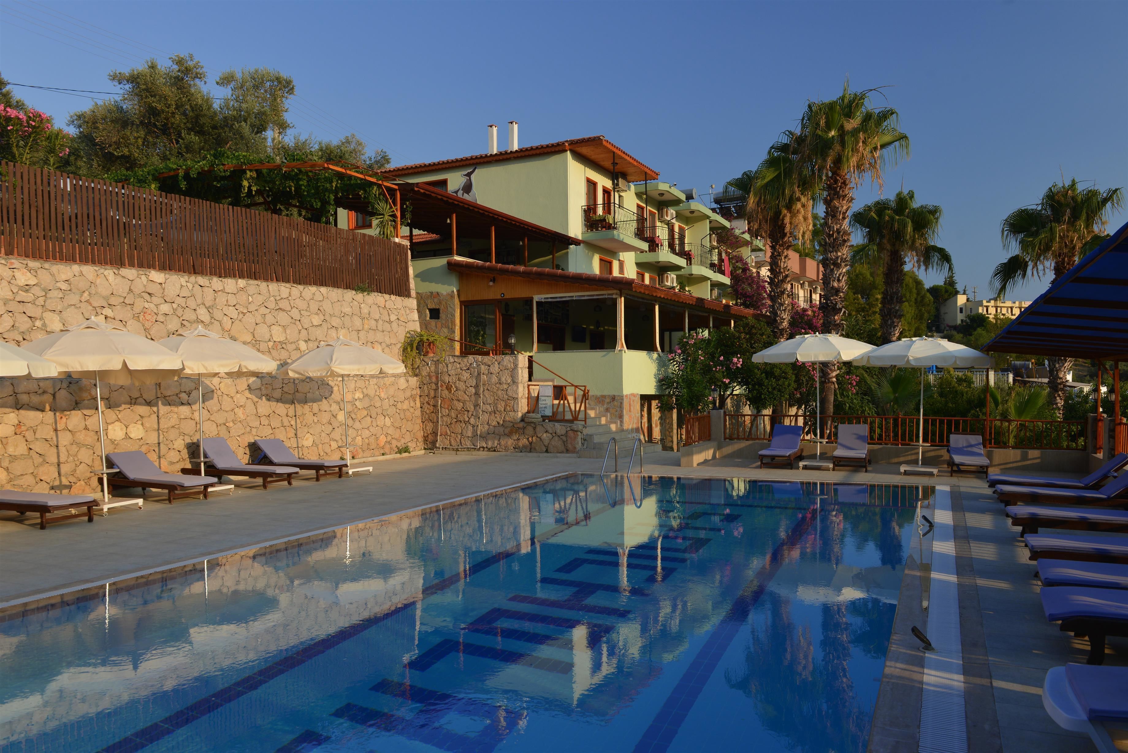 A swimming pool with lounge chairs and umbrellas, a stone wall, and a multi-story building with balconies.