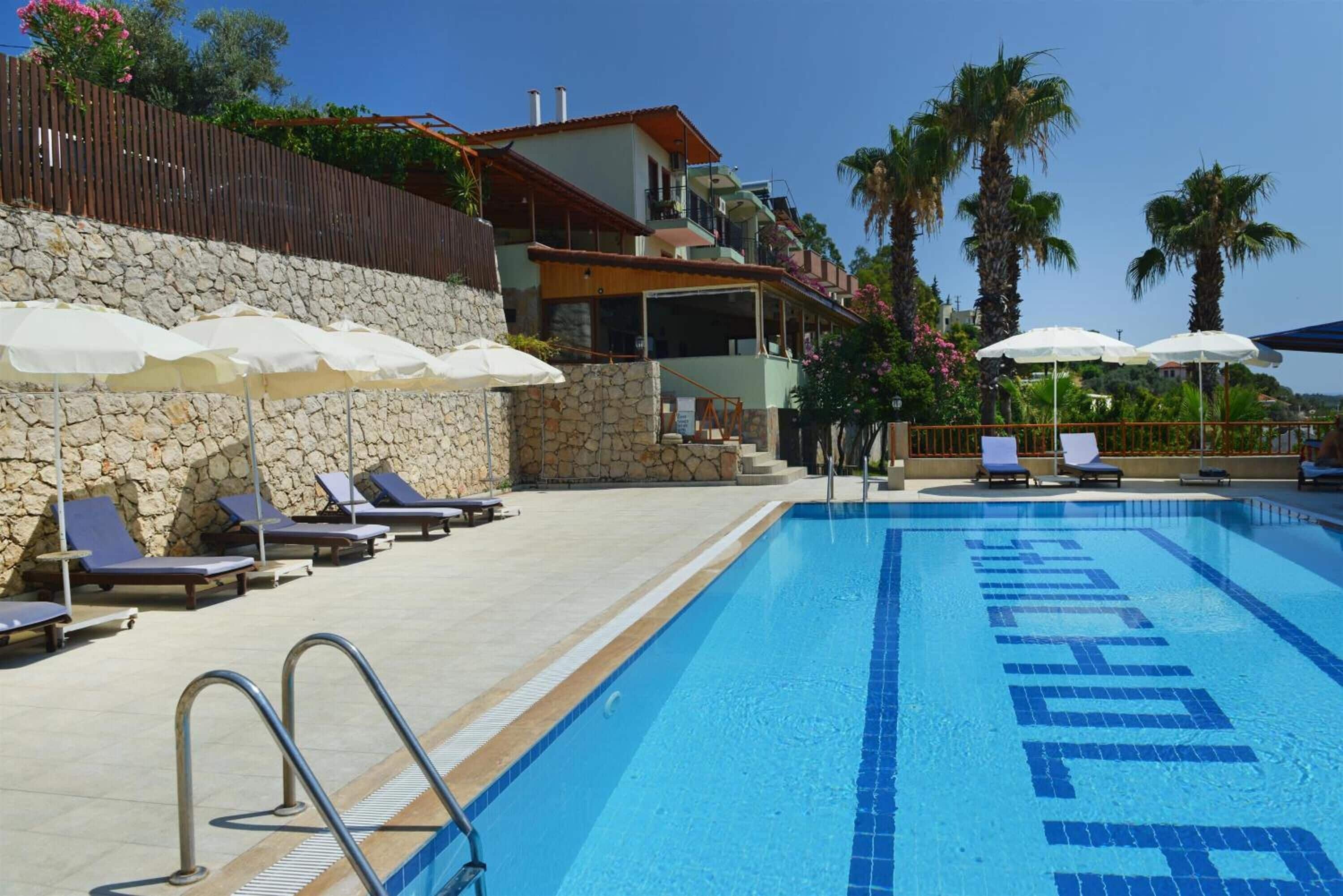 Una piscina con sillones de descanso y sombrillas, un muro de piedra y un edificio con balcones.
