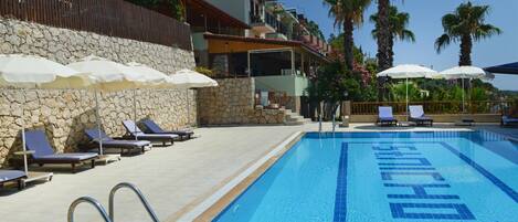 Una piscina con sillones de descanso y sombrillas, un muro de piedra y un edificio con balcones.