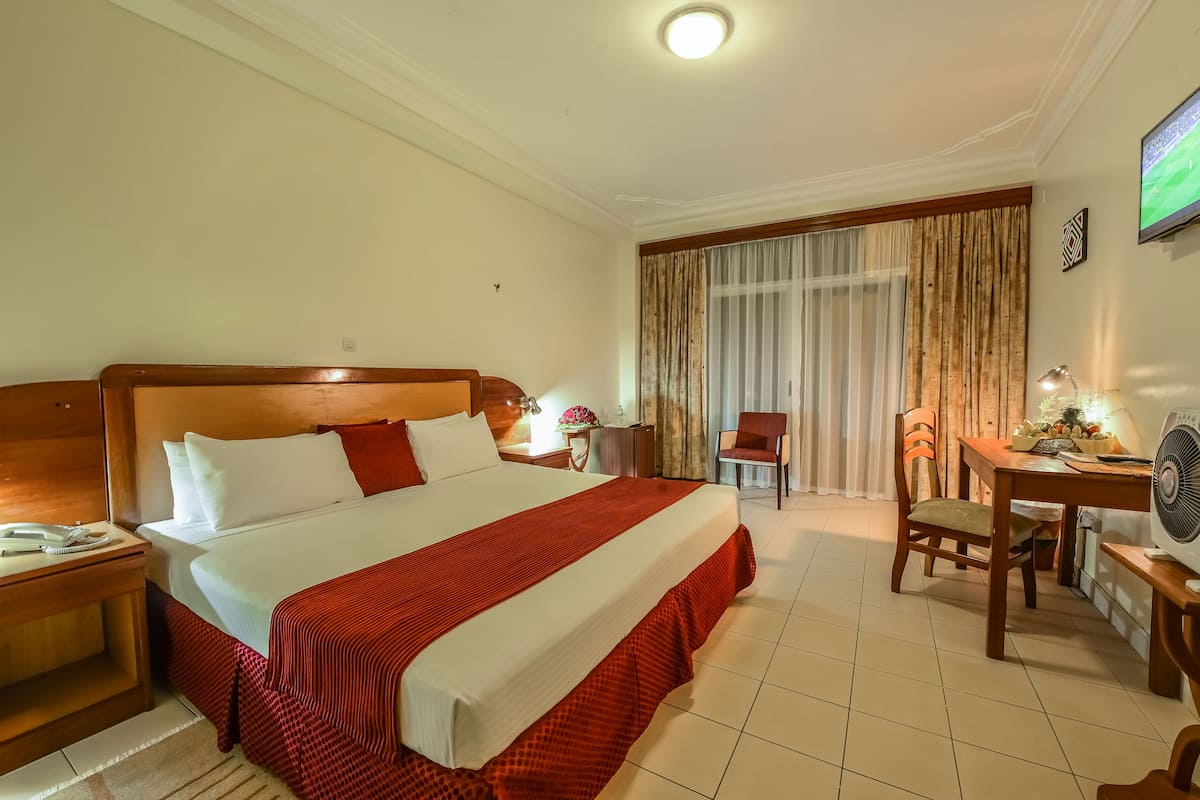 A hotel room with a large bed, a desk with a chair, a small table with a lamp, and a TV on the wall.